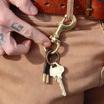 A person in tan pants holds the Flint Keychain by Tres Cuervos—featuring two keys and a handmade tassel—attached to their belt loop; "R G" tattoo is visible on their hand.