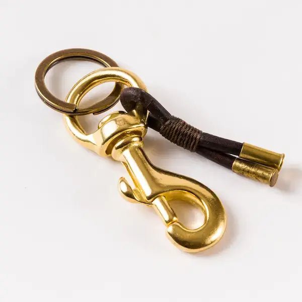 The Flint Keychain by Tres Cuervos features gold brass hardware, two rings, a clasp, and a dark cord with brass tips and a handmade tassel, all displayed on a white surface.