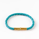 The Flint Single Waxed Canvas Bracelet by Tres Cuervos features a braided turquoise design and gold cylindrical clasp, shown here on a white background.