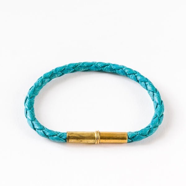 The Flint Single Waxed Canvas Bracelet by Tres Cuervos features braided turquoise canvas and a cylindrical brass clasp, shown on a white background.