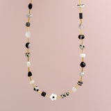 The Flower Beaded Necklace by Jill Makes features black, white, and gold beads with a delicate white flower accent, displayed on a light pink background. This elegant piece adds charm to any look.