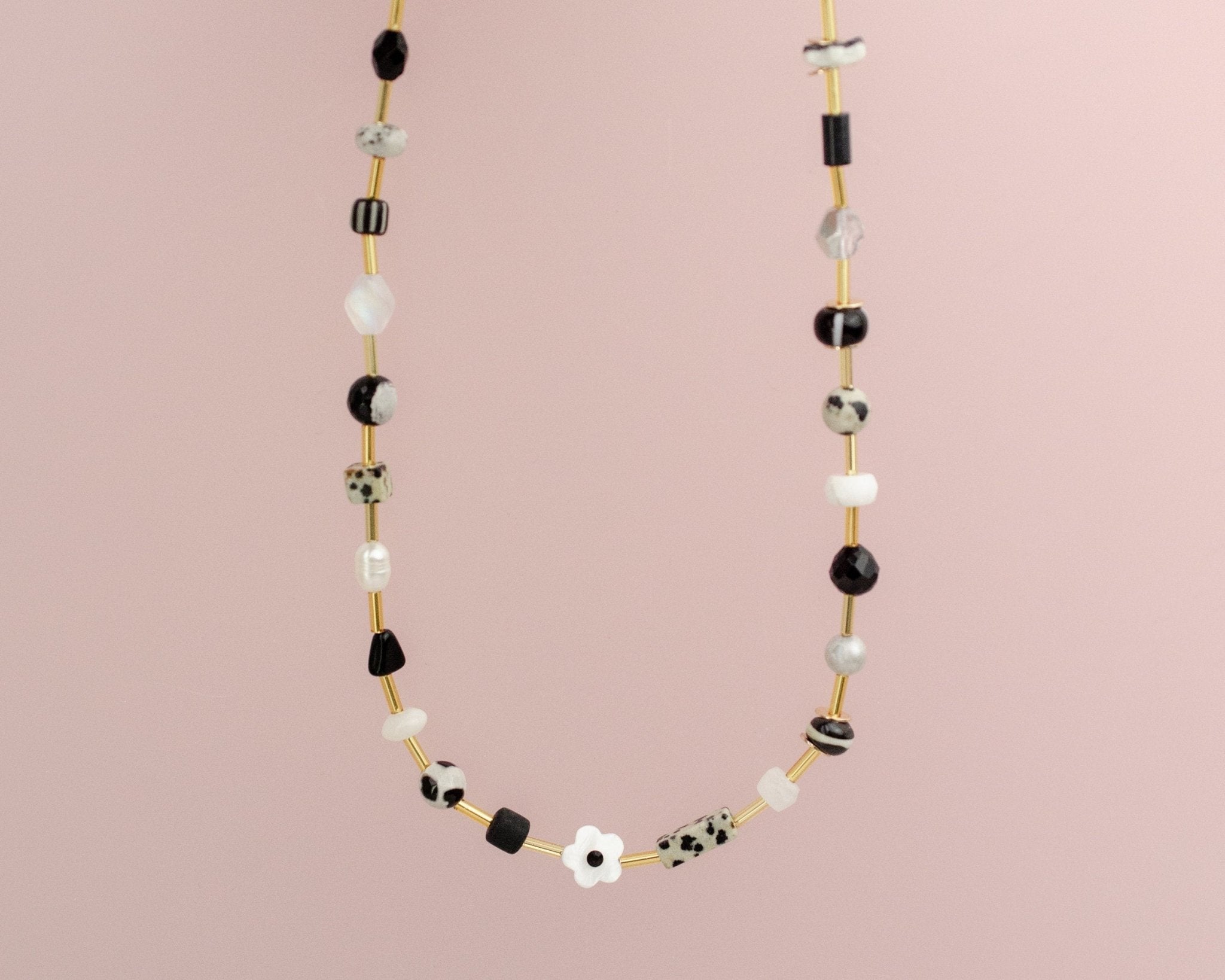 The Flower Beaded Necklace by Jill Makes features black, white, and gold beads with a delicate white flower accent, displayed on a light pink background. This elegant piece adds charm to any look.