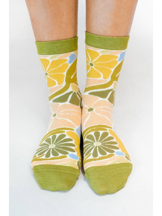 Model wearing the Flowers Knit Ankle Sock By Soulshine Co from Tailored Union, featuring a 70’s-inspired green, yellow, blue, and cream floral pattern, standing on a white surface.