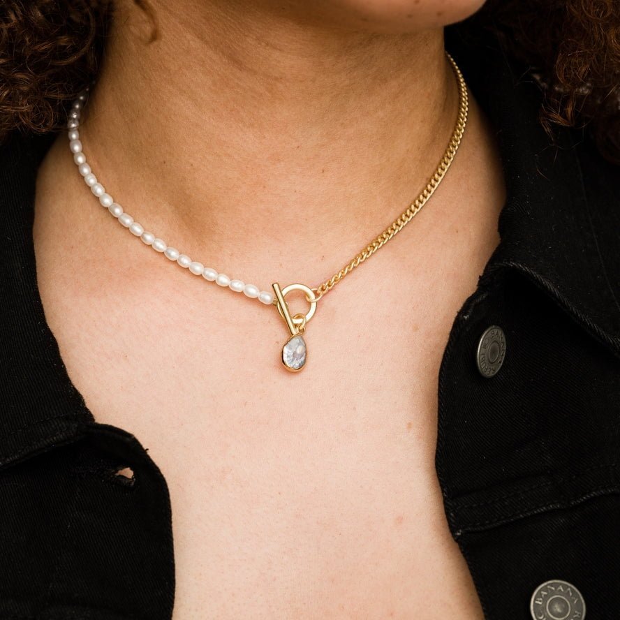 A person wearing the Larissa Loden Fonda Half Chain Pearl Necklace—featuring freshwater pearls, a gold chain, toggle clasp, and small pendant—over a black denim jacket. This hypoallergenic piece adds modern elegance to any outfit.