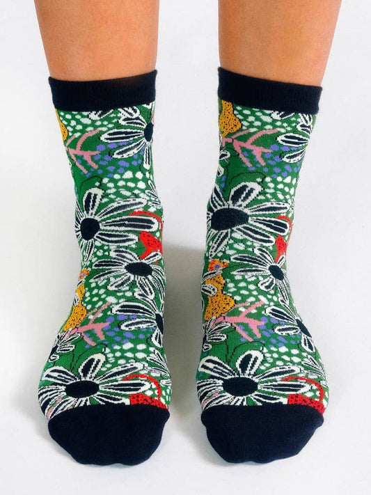 Person wearing Forest Knit Ankle socks by Tailored Union, featuring a vibrant floral pattern with daisies and leaves plus black toes, heels, and cuffs—an eye-catching design from the brand's colorful collection.