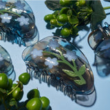 The Winona Irene Forget-Me-Not Light Blue Flower Acetate Hair Claw, featuring white and blue floral designs, is displayed on a light blue surface next to green artificial berries and leaves.