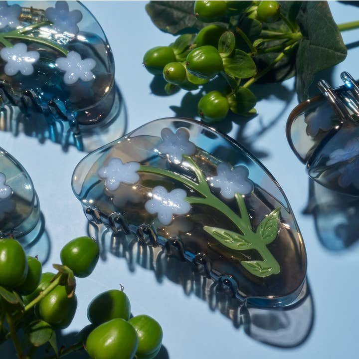 The Winona Irene Forget-Me-Not Light Blue Flower Acetate Hair Claw, featuring white and blue floral designs, is displayed on a light blue surface next to green artificial berries and leaves.