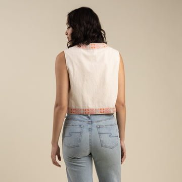 A woman faces away, wearing the Frontier Vest from Sendero Provisions Co.—a sleeveless beige top with floral embroidery and patterned trim in a cropped boxy fit—paired with light blue jeans against a plain background.