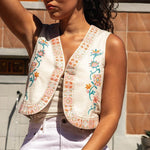 A woman stands outdoors in sunlight wearing the Frontier Vest by Sendero Provisions Co., a cream-colored embroidered floral vest with a cropped boxy fit, layered over a white top and pants.