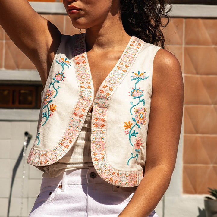 A woman stands outdoors in sunlight wearing the Frontier Vest by Sendero Provisions Co.—a sleeveless cream vest with colorful floral embroidery—styled over a white top and high-waisted pants.
