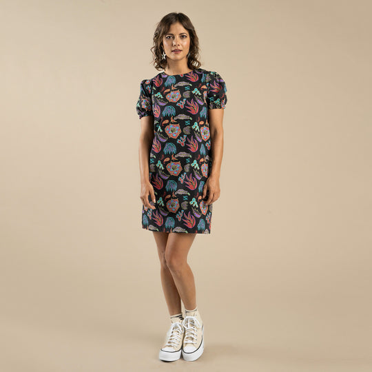 A woman stands against a beige background, wearing the Gabriela Shift Dress by Sendero Provisions Co.—a short-sleeve, knee-length organic cotton dress with a colorful floral print—paired with white high-top sneakers.