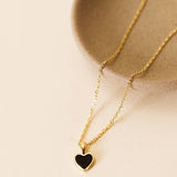 The Gemstone Heart Pendant Necklace - Black Tourmaline by JaxKelly, a tarnish-free gold chain with a black heart pendant, rests beside a beige ceramic dish on a light surface.