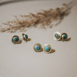 Three pairs of Gibbon Studs by CommonForm, featuring round turquoise stones set in brushed brass, are arranged on a light surface with dried grass accents in the background.