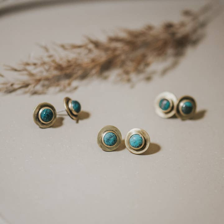 Three pairs of Gibbon Studs by CommonForm, featuring round turquoise stones set in brushed brass, are arranged on a light surface with dried grass accents in the background.