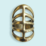The Gladiator Ring by Ornamental Things features a wide, openwork design with three horizontal bars crossed by a central vertical bar, crafted from 24kt gold-plated brass and shown against a light blue background.