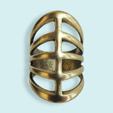 The Gladiator Ring by Ornamental Things features a wide, openwork design with three horizontal bars crossed by a central vertical bar, crafted from 24kt gold-plated brass and shown against a light blue background.