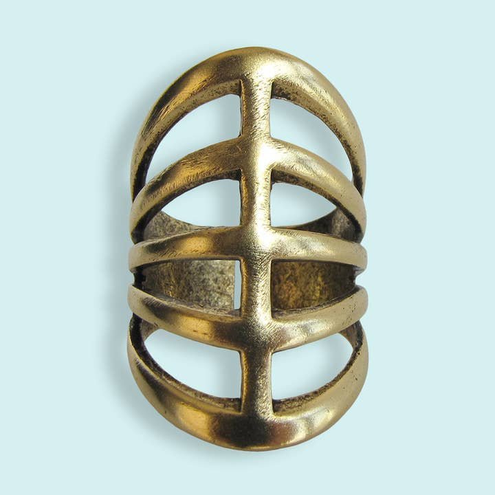 The Gladiator Ring by Ornamental Things features a wide, openwork design with three horizontal bars crossed by a central vertical bar, crafted from 24kt gold-plated brass and shown against a light blue background.