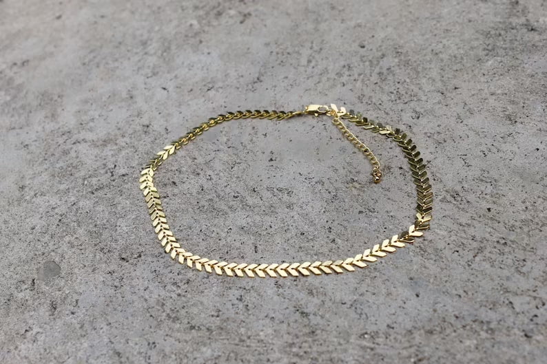 The Gold Chevron Choker by Vintage Acorn, 16k gold plated with an adjustable clasp, is displayed on a textured gray surface.