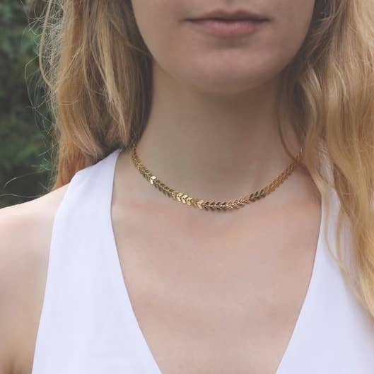A woman wears the Gold Chevron Choker by Vintage Acorn with a white top, shown from the shoulders up against a blurred outdoor background.