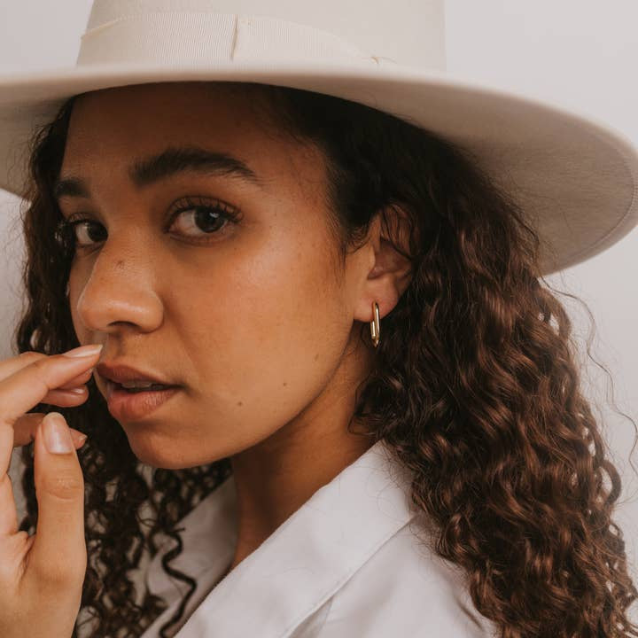 A woman with curly hair wears a wide-brimmed white hat, a white shirt, and JaxKelly Gold Hoop - Large Rectangle Earrings, looking at the camera and touching her face.