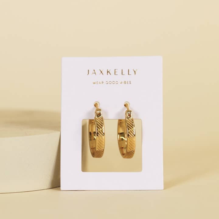 A pair of tarnish-free textured gold hoop earrings from JaxKelly displayed on a white card with the brand name and slogan "Wear Good Vibes.