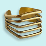 A gold cuff bracelet with four parallel, angular bands set against a light blue background, paired perfectly with the Gold Line Ring by Ornamental Things for an elegant touch.