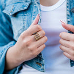 A person wearing the Gold Line Ring by Ornamental Things, a white shirt, and a light blue denim jacket, holding the jacket with both hands.