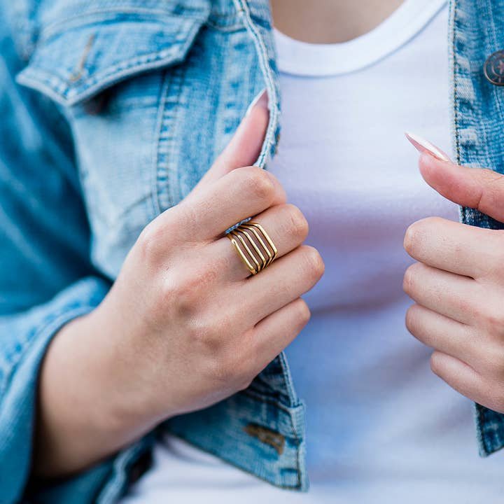 A person wearing the Gold Line Ring by Ornamental Things, a white shirt, and a light blue denim jacket, holding the jacket with both hands.