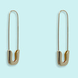 Gold Safety Pin Earrings by Ornamental Things—oversized, nickel free hoops shaped like safety pins. Stylish and displayed on a light blue background.