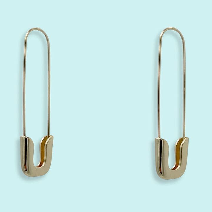 Gold Safety Pin Earrings by Ornamental Things—oversized, nickel free hoops shaped like safety pins. Stylish and displayed on a light blue background.