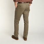 A man stands with his back to the camera, hands in pockets, wearing Duck Head Gold School Chino pants in olive green, a brown plaid shirt, brown belt, and brown shoes.