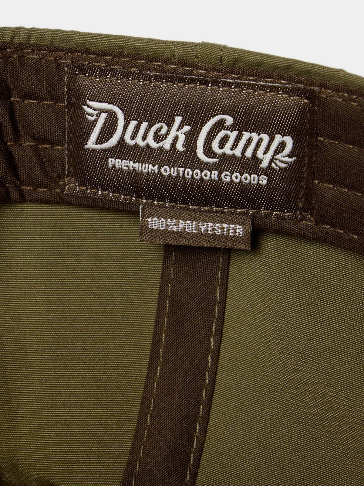 Close-up of a Duck Camp clothing label reading "Duck Camp Premium Outdoor Goods" and "100% Polyester" on green and brown fabric, featured on the Grandpa Hat | Gator by Duck Camp—ideal for your next outdoor adventure.