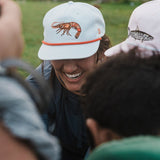 Someone smiles outdoors, wearing the Duck Camp Grandpa Hat Gulf Shrimp snapback with a shrimp illustration. The adjustable fit ensures comfort all day.