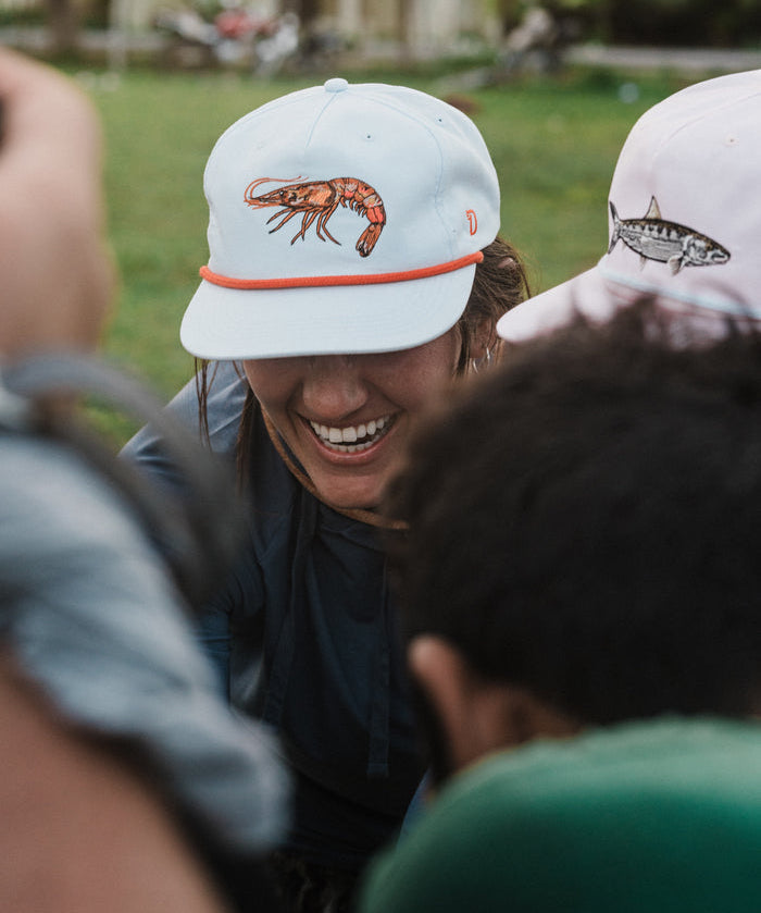 Someone smiles outdoors, wearing the Duck Camp Grandpa Hat Gulf Shrimp snapback with a shrimp illustration. The adjustable fit ensures comfort all day.