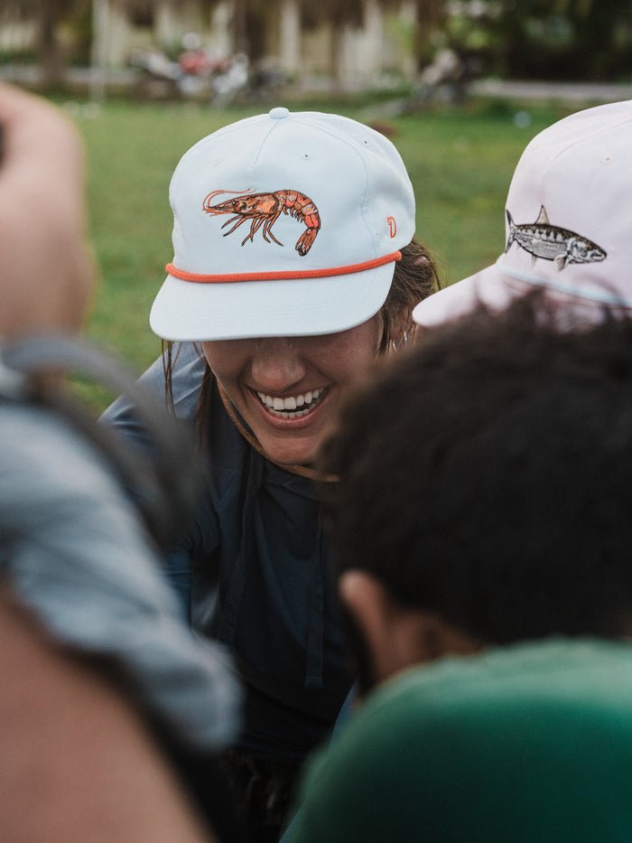 Someone smiles outdoors, wearing the Duck Camp Grandpa Hat Gulf Shrimp snapback with a shrimp illustration. The adjustable fit ensures comfort all day.