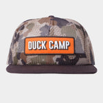 The Gulf Trucker | Wetland by Duck Camp is a camo flat-brim hat with an orange "DUCK CAMP" patch in white. It features breathable mesh panels for comfort and airflow, making it ideal for outdoor adventures.