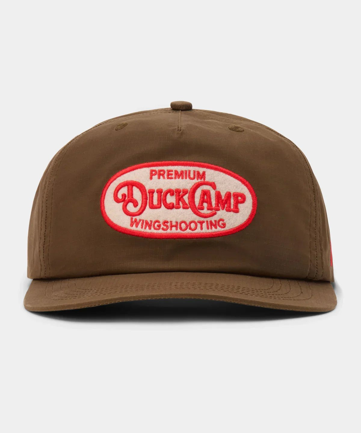 The Gunnison Wingshooting Badge Waxed Hat by Duck Camp is a brown, water-resistant canvas cap featuring a snapback closure and a red and white oval patch with "Premium Duck Camp Wingshooting" on the front.