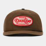 The Gunnison Wingshooting Badge Waxed Hat by Duck Camp is a brown, water-resistant canvas cap featuring a snapback closure and a red and white oval patch with "Premium Duck Camp Wingshooting" on the front.