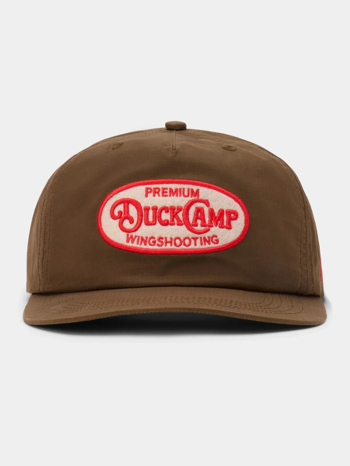 The Gunnison Wingshooting Badge Waxed Hat by Duck Camp is a brown, water-resistant canvas cap featuring a snapback closure and a red and white oval patch with "Premium Duck Camp Wingshooting" on the front.