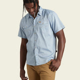 A man wears the Howler Brothers H Bar B Snapshirt in Faded Blue Oxford with chest pockets and pearl snaps, paired with tan pants, standing against a plain light background.