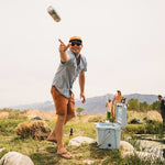 A man in sunglasses and an orange cap, wearing the H Bar B Snapshirt in Faded Blue Oxford by Howler Brothers, tosses a can toward a cooler outdoors with mountains and people in the background.