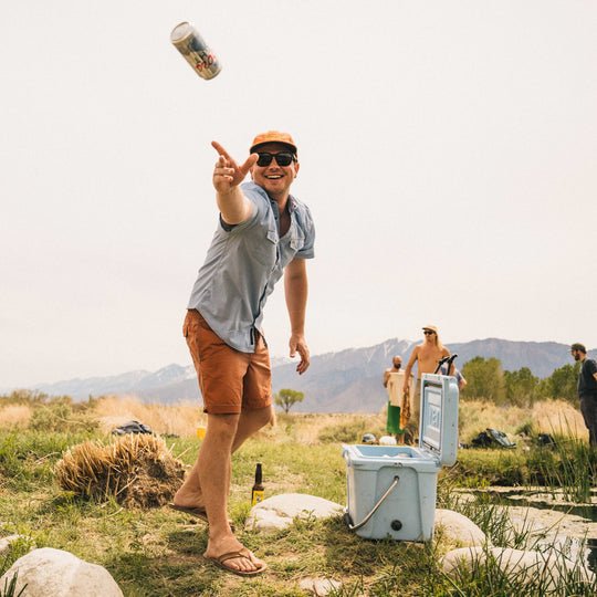 A man in sunglasses wearing the Howler Brothers H Bar B Snapshirt in Faded Blue Oxford tosses an empty can toward a cooler outdoors, with mountains and people in the background.