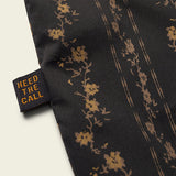 Close-up of the H Bar B Snapshirt Hibiscus Ribbons by Howler Brothers, showing black fabric with a gold floral pattern and a "HEED THE CALL" tag in yellow text, highlighting its refined, classic style.