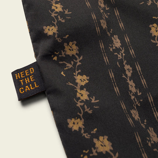 Close-up of the H Bar B Snapshirt Hibiscus Ribbons by Howler Brothers, showing black fabric with a gold floral pattern and a "HEED THE CALL" tag in yellow text, highlighting its refined, classic style.