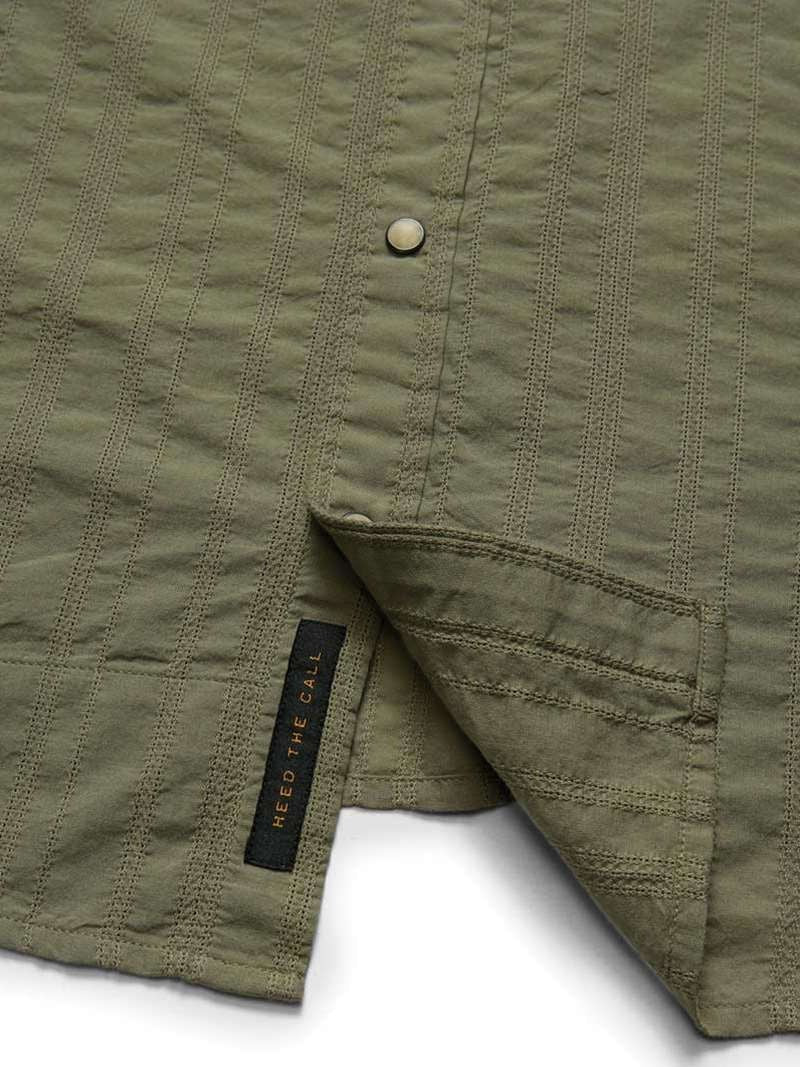 Close-up of the olive green H Bar B Snapshirt by Howler Brothers, featuring textured fabric, pearl snaps, and a visible black "HEED THE CALL" tag near the hem.