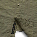 Close-up of the olive green H Bar B Snapshirt by Howler Brothers, featuring textured fabric, pearl snaps, and a visible black "HEED THE CALL" tag near the hem.