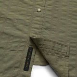 Close-up of the olive green H Bar B Snapshirt by Howler Brothers, featuring textured fabric, pearl snaps, and a visible black "HEED THE CALL" tag near the hem.