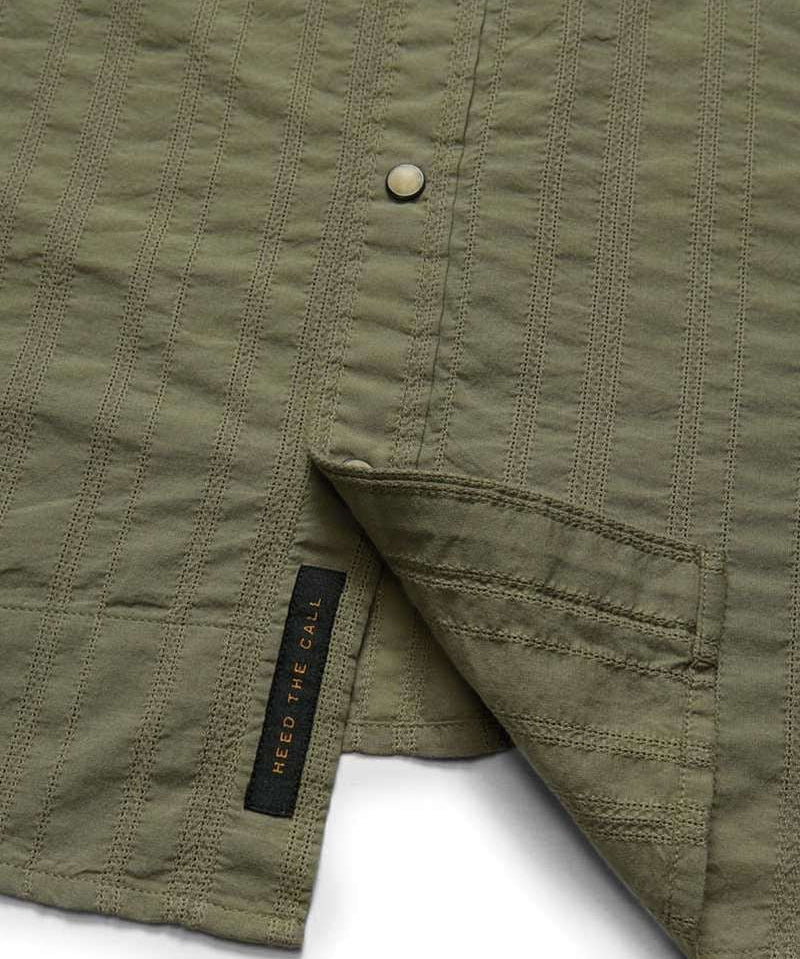 Close-up of the olive green H Bar B Snapshirt by Howler Brothers, featuring textured fabric, pearl snaps, and a visible black "HEED THE CALL" tag near the hem.