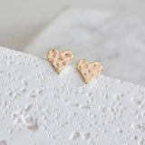 A pair of small, 18K gold plated Hammered Heart Stud Earrings from Mesa Blue with a textured finish, shown on a white surface. Lightweight and elegant, these Mesa Blue earrings enhance any outfit.