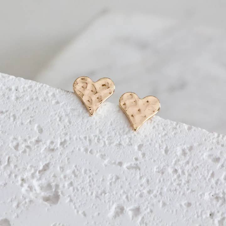 A pair of small, 18K gold plated Hammered Heart Stud Earrings from Mesa Blue with a textured finish, shown on a white surface. Lightweight and elegant, these Mesa Blue earrings enhance any outfit.